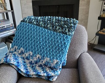 Könnte beinhalten: Eine blaue, weiße und grüne gehäkelte Wolldecke mit einem strukturierten, bauschigen Design. Die Decke liegt über einem grauen Sessel.