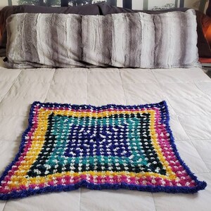 May include: A vibrant, square crochet blanket with a textured design. The blanket features a palette of blue, teal, yellow, pink, and white. The blanket is placed on a bed with a white duvet and a grey pillow.