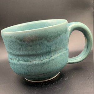 Pode incluir: Uma caneca de cerâmica azul-esverdeada com uma base larga e arredondada e uma borda ligeiramente alargada. A caneca tem um acabamento texturizado e envidraçado com variações sutis de cor.