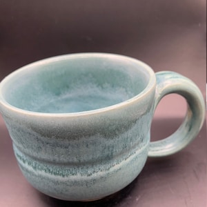 Puede incluir: Una taza de cerámica hecha a mano con un esmalte turquesa. La taza tiene un tazón ancho y redondeado y una asa robusta.