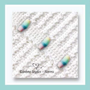May include: Three small, barrel-shaped toggles featuring a gradient of pastel colours including yellow, green, blue, and pink. The toggles are arranged on a white, textured, knitted fabric background. The text Rainbow Toggles - Aurora is visible.