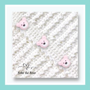 May include: Three pink bear-shaped buttons with black eyes and noses are scattered on a white, textured fabric. The text "Bobo the Bear" is printed in black below a heart-shaped design.
