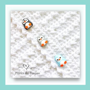 May include: Three penguin-shaped embellishments in black, grey, and blue with orange feet and beaks are scattered on a white, textured surface. The text "Pebbles the Penguin" is at the bottom. These are likely decorative craft supplies.