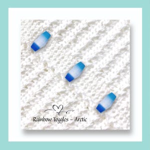 May include: Three small toggle fasteners with a gradient colour transition from white to blue, positioned on a white knitted fabric. The text "Rainbow Toggles - Arctic" is printed at the bottom with a small heart icon above the word Rainbow.