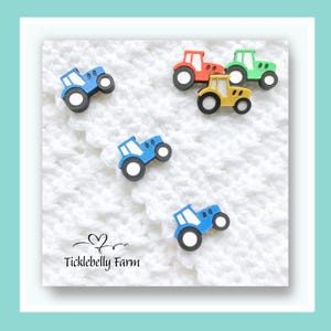 May include: A selection of colourful tractor-shaped embellishments in red, green, yellow, and blue. The tractors have black wheels and white accents. The background is a white, textured fabric. The text "Ticklebelly Farm" is visible.