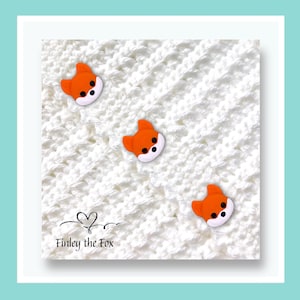 May include: A white crocheted blanket adorned with three orange fox-shaped buttons. Each button features a white face and black eyes. The text "Finley the Fox" is written in black script at the bottom.