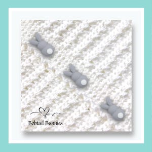 May include: A white crocheted blanket with three grey bunny figures with white tails. The blanket has a textured pattern. The text "Bobtail Bunnies" is at the bottom left corner of the image.