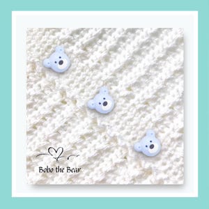 May include: A white crocheted blanket with three light blue bear-shaped buttons. The buttons have black eyes and noses. The text "Bobo the Bear" is at the bottom of the blanket.