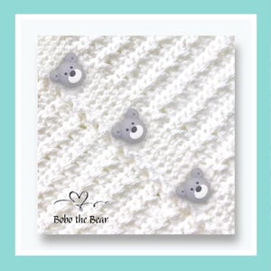 May include: A white crocheted blanket with three grey bear-shaped buttons. The buttons have white faces and black eyes. The text "Bobo the Bear" is at the bottom left corner of the blanket.