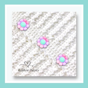 May include: A close-up of white crocheted fabric featuring three pastel flower decorations. The flowers are pink, purple, and mint green. The text "Rainbow Daisies" is written in a script font below a heart symbol.