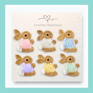 May include: Six crochet bunny appliques arranged in two rows on a white card labelled Crochet Applique. Each bunny features a tan head with a black eye, long ears, and a small white tail, wearing a jumper in pink, white, blue, purple, yellow, or green.