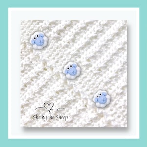 May include: A white crocheted blanket featuring three sheep embellishments. The sheep have blue faces and white wool. The blanket is branded with the text "Shelby the Sheep" in black.