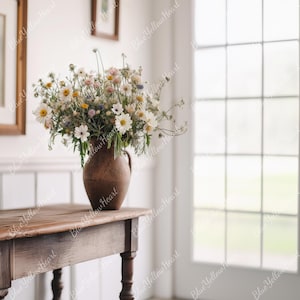 May include: A rustic wooden table with a vase of white flowers in a brown ceramic vase. The table is in front of a window with white trim and glass panes.