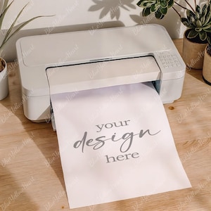 May include: A white printer with a sheet of paper emerging from the output tray. The paper has the words "your design here" printed on it in a stylish font. The printer is on a light wood surface.