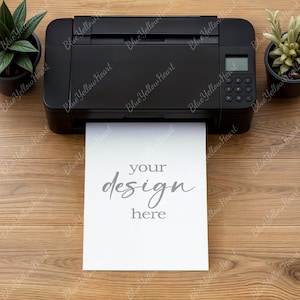 May include: A black printer with a digital display and control buttons is printing a white sheet of paper. The paper has the words "your design here" printed on it. The printer is on a wooden surface, with small potted plants on either side.