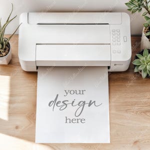 May include: A white printer with a sheet of paper emerging, displaying the words "your design here" in gray script. The printer sits on a light wood surface, with potted plants on either side.