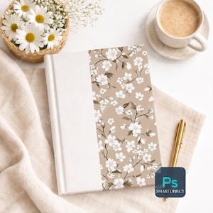 May include: A white journal with a floral design on a tan background. The floral pattern features white flowers and brown stems. A cup of coffee, a gold pen, and a basket of daisies are also in the image.