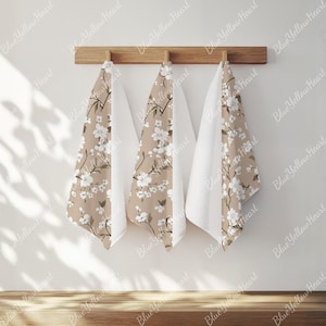 May include: Three decorative towels hanging on a wooden rack. Two towels feature a beige floral pattern with white blossoms and dark stems. The center towel is solid white. The towels are hanging against a white wall.