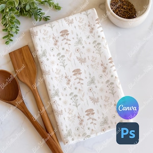 May include: A folded white kitchen towel with a floral pattern in shades of brown and green. Two wooden spoons and a wooden spatula are next to the towel. A small bowl of seeds and some greenery are also in the image.