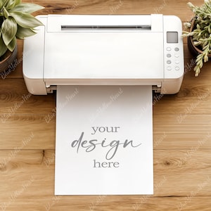 May include: A white printer with a sheet of paper emerging, displaying the words "your design here" in gray script. The printer is on a wooden surface, with potted plants on either side.