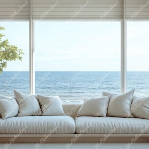 May include: A white couch with multiple pillows sits in front of a large window with a view of the ocean and a beach. The couch is made of a soft, white fabric and has a simple, modern design.