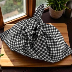 May include: A black and white checkered fabric wrapping a package, tied in a knot. The fabric is on a wooden cutting board, with a window and plants in the background. The pattern is a classic gingham.