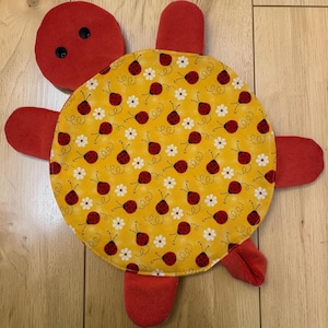 May include: A turtle-shaped fabric toy featuring a bright yellow shell patterned with small red ladybugs and white flowers. The head, four flippers, and tail are made of solid red fabric, with two black buttons serving as eyes on the head.