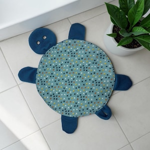 May include: A turtle-shaped fabric mat featuring a circular shell with a teal background and a repeating pattern of small dark blue and yellow cross stitches. The head, four flippers, and tail are made of solid dark blue fabric.