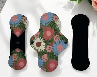 Puede incluir: Tres compresas menstruales de tela reutilizables con estampado floral. Las compresas son de diferentes tamaños y formas. La compresa más grande es de color verde claro con un estampado floral rosa, blanco y azul. La compresa mediana es de color verde oscuro con un estampado floral rosa, blanco y azul. La compresa más pequeña es de color negro.