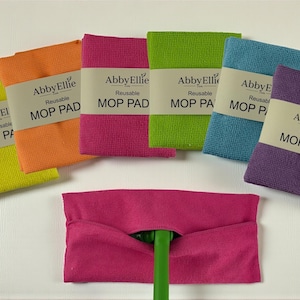 Puede incluir: Un conjunto de mopa reutilizables de colores brillantes: amarillo, naranja, rosa, verde, azul y morado. Cada mopa lleva la inscripción "Abby Ellie Reusable MOP PAD" o "MOP PADS". Una mopa rosa está sujeta a un mango verde.