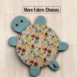 May include: A turtle-shaped fabric item with a floral patterned shell featuring red, yellow, and blue flowers on a white background. The head, limbs, and tail are solid light blue. The head has two black button eyes. Text reads: More Fabric Choices.