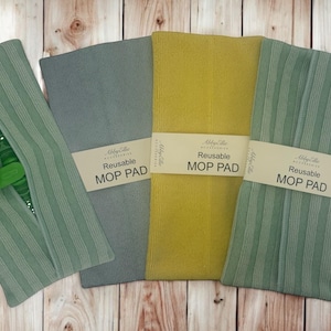 Puede incluir: Cuatro almohadillas de fregona reutilizables en tonos verdes, grises y amarillos se muestran sobre una superficie de madera. Cada almohadilla está envuelta con una etiqueta blanca que dice "Reusable MOP PAD". Una almohadilla está unida a un mango de fregona verde.