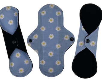 Puede incluir: Tres compresas menstruales de tela reutilizables con fondo azul y estampado de margaritas blancas. Las compresas están dobladas por la mitad y tienen tela negra en la parte posterior con un cierre de presión.