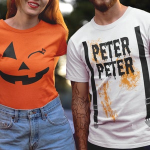 May include: Two people wearing matching Halloween costumes. The person on the left is wearing an orange t-shirt with a black winking jack-o-lantern face. The person on the right is wearing a white t-shirt with the words "Peter Peter" in black dripping letters and black suspenders.