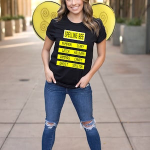 May include: A woman wearing a black t-shirt with the words "Spelling Bee" and a list of Halloween words in yellow boxes. She is wearing blue jeans and white sneakers. She is also wearing yellow bee wings.