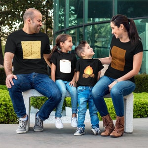 May include: A family of four wearing matching black t-shirts with camping-themed graphics. The shirts feature a graham cracker, marshmallow, campfire, and chocolate bar design. They are sitting on a concrete bench, with a green background.