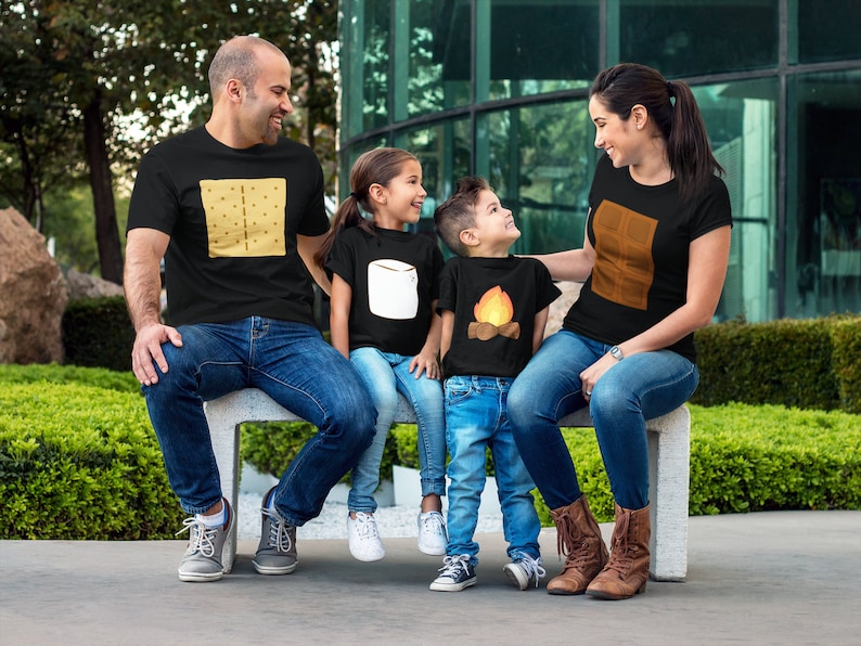 May include: A family of four wearing matching black t-shirts with camping-themed graphics. The shirts feature a graham cracker, marshmallow, campfire, and chocolate bar design. They are sitting on a concrete bench, with a green background.