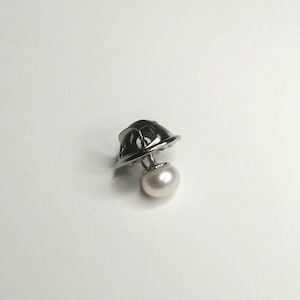 May include: A silver-toned lapel pin featuring a small, stylized helmet design with a dangling white pearl. The pin is set against a plain white background, highlighting the jewelry's details and craftsmanship.