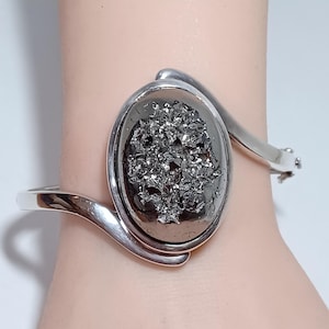 May include: A silver-toned cuff bracelet featuring an oval, dark gray, textured stone. The bracelet has a hinged clasp and a sleek, modern design. The stone is the focal point, set in a polished silver frame.