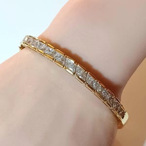 May include: A gold bracelet with a row of square-cut, clear gemstones. The bracelet is set in a gold metal band, creating a continuous line of sparkle. The clasp is visible on the left side of the bracelet.
