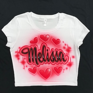 May include: White crop top with a red and black airbrushed design. The design features the name "Melissa" in black script with red hearts and sparkles around it.