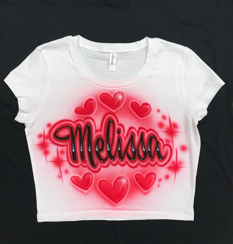 May include: White crop top with a red and black airbrushed design. The design features the name "Melissa" in black script with red hearts and sparkles around it.