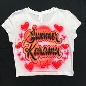May include: White crop top with a red, orange, and black airbrushed design. The design features the name "Summer Karamu" with the year "'24" below it. The design is surrounded by red hearts and sparkling stars.