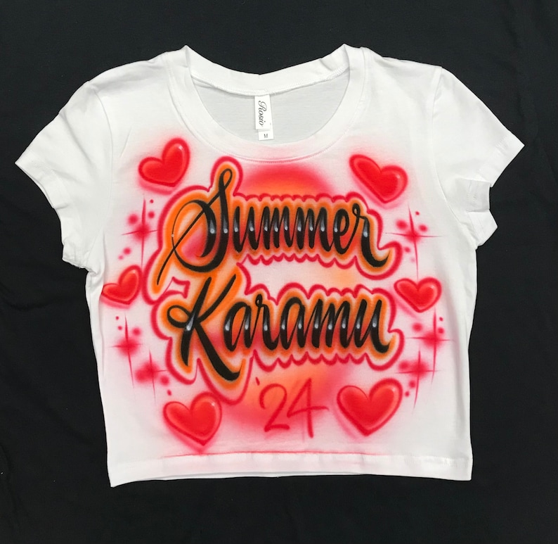 May include: White crop top with a red, orange, and black airbrushed design. The design features the name "Summer Karamu" with the year "'24" below it. The design is surrounded by red hearts and sparkling stars.