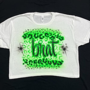 May include: White cropped t-shirt with a green and black leopard print design. The text "Success is a brat" is written in black on the shirt.