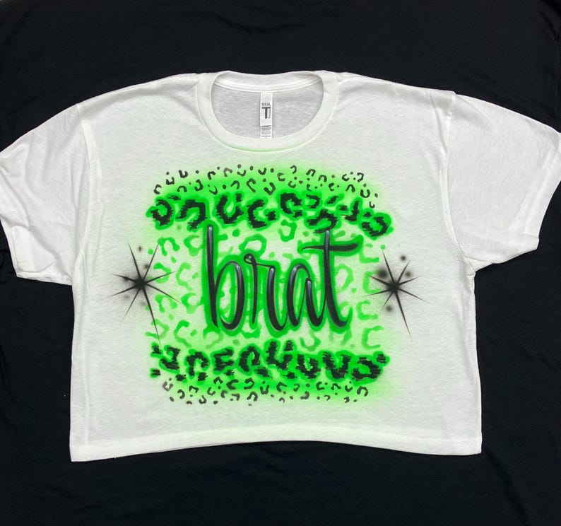 May include: White cropped t-shirt with a green and black leopard print design. The text "Success is a brat" is written in black on the shirt.