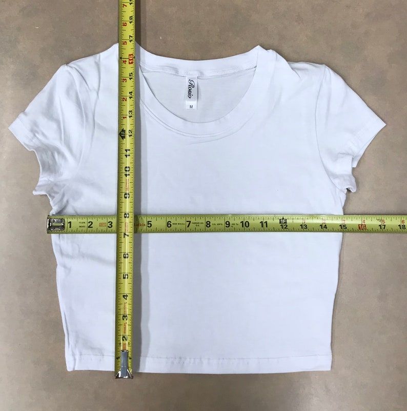 May include: White cropped t-shirt with short sleeves and a round neckline. The shirt has a tag that says "Rosio" and "M". The shirt is measured with a tape measure, showing a width of 17 inches and a length of 15 inches.