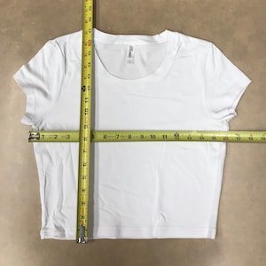 May include: White cropped t-shirt with short sleeves. The shirt has a round neckline and a tag that says "Rosio" on the front. The shirt is measured with a tape measure, showing the length and width.