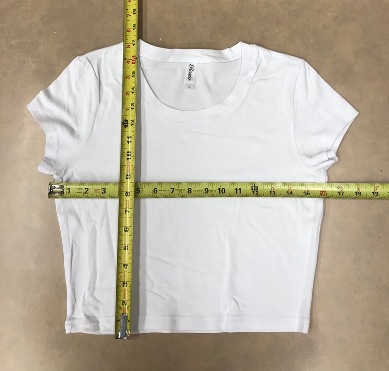 May include: White cropped t-shirt with short sleeves. The shirt has a round neckline and a tag that says "Rosio" on the front. The shirt is measured with a tape measure, showing the length and width.