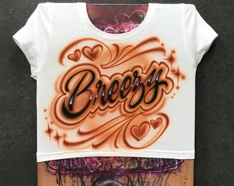 May include: White crop top with the word "Breezy" in a brown and black airbrushed design. The design includes hearts and swirls. The shirt has a round neckline and short sleeves.